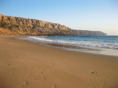 Huzurlu kumlu bir sahil. Arka planda muhteşem bir uçurum yükseliyor. Doğanın manzarasını güzelleştiriyor.