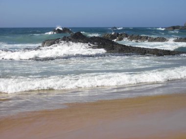 Dalgalar yavaşça kumlu bir plaja çarpıyor ve arka planda kayalar belirgin bir şekilde ortaya çıkıyor. Çarpıcı bir doğal manzara yaratıyor.