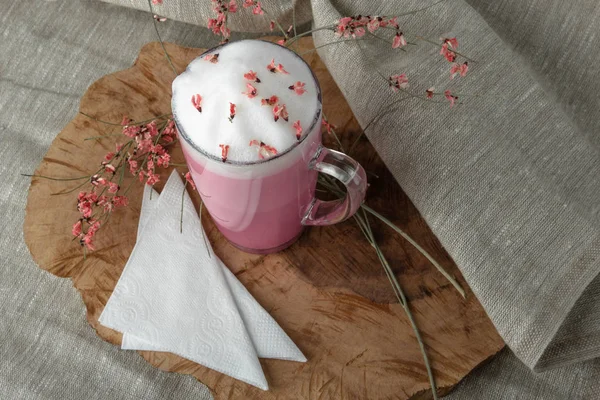 Pink coffee latte macchiato in a glass cup on a wooden backing decorated with dried flowers with air foam