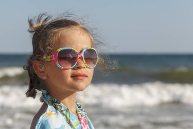 Güneş gözlüğü takan küçük kız güneşi deniz arka planına yansıtıyor