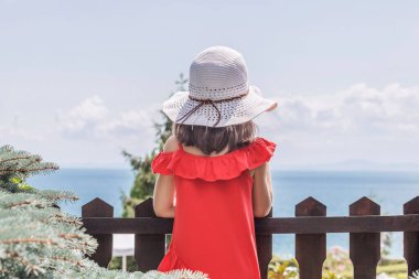 Beyaz plaj şapkalı ve kırmızı elbiseli küçük bir kız sırtı kameraya dönük duruyor ve denize ve bulutlu gökyüzüne bakıyor. Denizden gelen prensi bekliyorum..