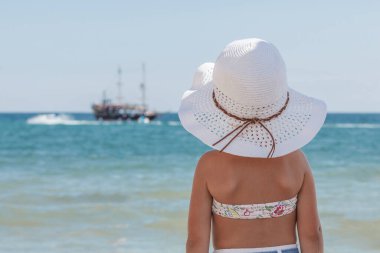 Bikinili ve beyaz plaj şapkalı küçük bir kız kumlu bir plajda oturuyor sırtı kameraya dönük denize bakıyor ve yatı uzaklarda seyrediyor.