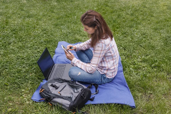 Workplace on a blue rug in nature. Woman sitting in the garden near the laptop with a marker in her hand and looking at the phone