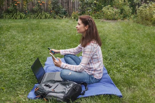 Workplace on a blue rug in nature. Woman sitting in the garden near the laptop and thinking about something with a marker and a phone in hand