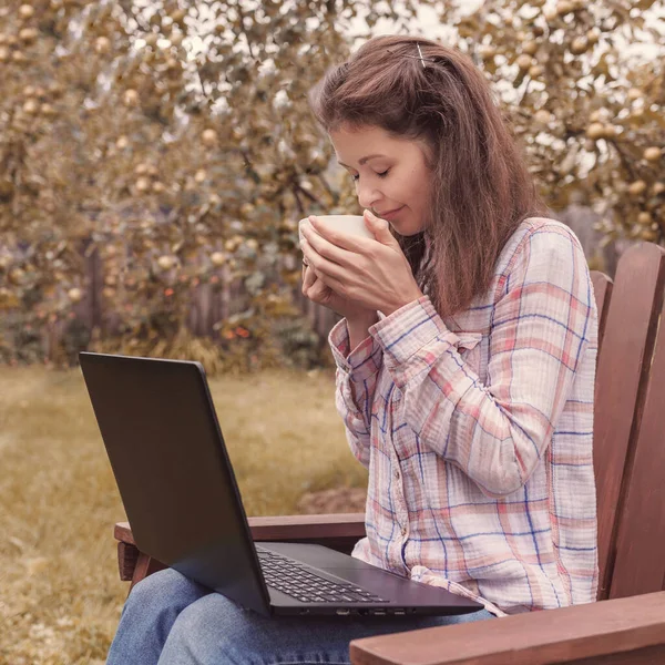 Young woman sitting in the garden at the laptop with a cup of tea in her hands