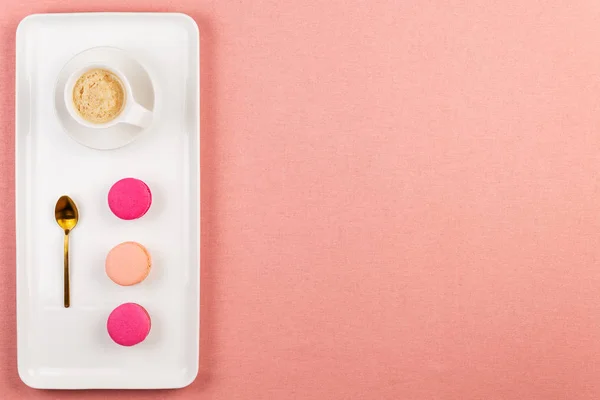 Różowe francuskie makaroniki lub makaroniki, filiżankę kawy i złotą łyżkę na prostokątnych białych płytce na różowym obrus tle z kopią przestrzeni. — Zdjęcie stockowe