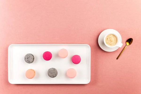 Macarons ou macarons français roses et gris, sur une assiette rectangle blanche avec tasse à café et cuillère dorée sur un fond de nappe rose avec espace de copie . — Photo