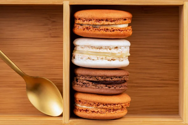 Brown and white french macarons or macaroons, (chocolate, coffee, toffee, salted caramel and vanilla) stacked in a bamboo box with a golden tea spoon and empty space. — Stock Photo, Image