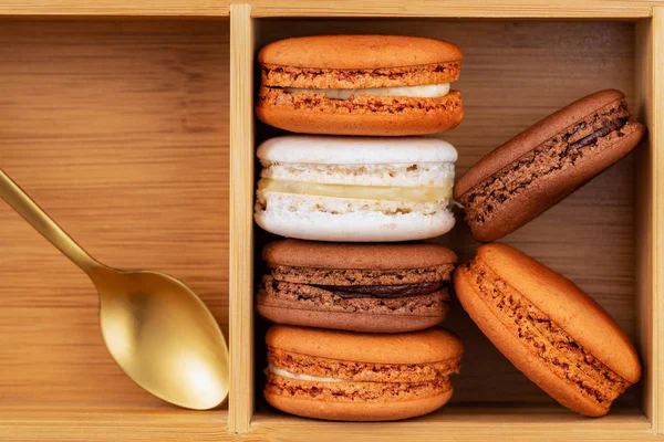 Brown and white french macarons or macaroons, (chocolate, coffee, toffee, salted caramel and vanilla) stacked in a bamboo box with a golden tea spoon. — Stock Photo, Image