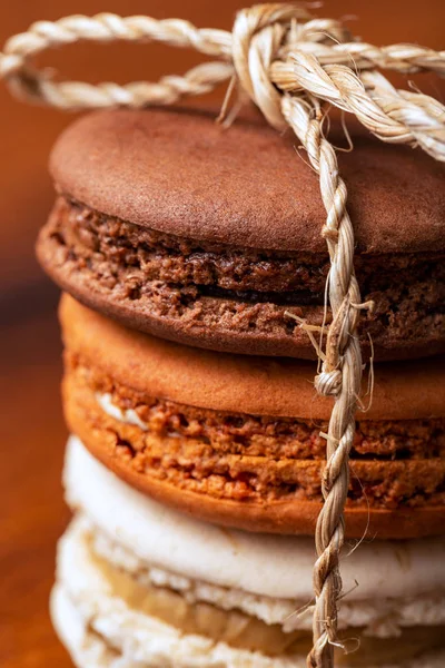 Three brown and white french macarons or macaroons, (chocolate, salted caramel and vanilla) tighten together with a natural string over a vintage wood background. Close-up. — Stock Photo, Image