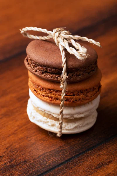 Three brown and white french macarons or macaroons, (chocolate, salted caramel and vanilla) tighten together with a natural string over a vintage wood background. — Stock Photo, Image