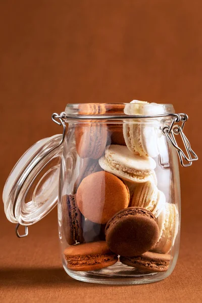 Brown and white french macarons or macaroons, stacked inside an open vintage glass jar over a chocolate brown background in vertical view. — Stock Photo, Image