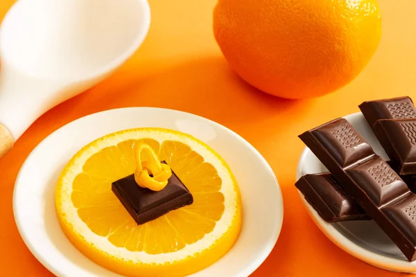 Chocolate pieces over orange slice, full orange and white cooking spoon over orange background.