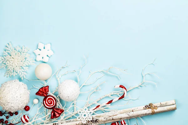 Frost like white and red christmas composition with branches, birch sticks, white glass spheres, decoration sweets, candy cane and snow flakes over a light blue background. Flat lay, top view with copy space.