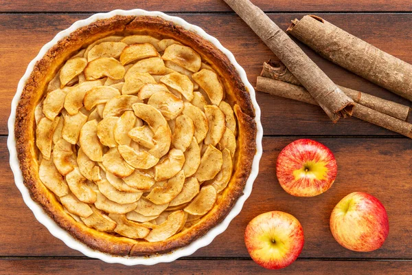 Homemade baked French apple tart, an open faced apple pie, in a baking white ceramic dish aside Gala red apples and cinnamon sticks, all on a vintage dark wood background. Flat lay, top view.