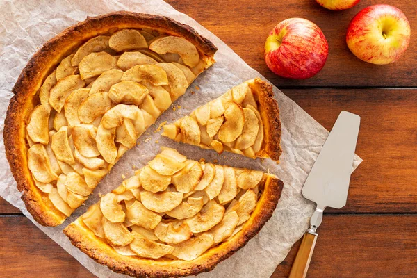 Homemade baked French apple tart, an open faced apple pie, aside Gala apples and silver vintage cake server or tart server on a baking paper sheet on vintage wood table. Flat Lay, top view.