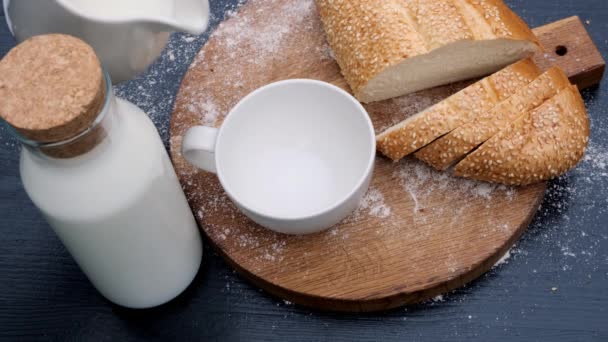 Großaufnahme des Einfüllens von Milch in weißen Becher in einem hölzernen Hintergrund neben Brot. — Stockvideo