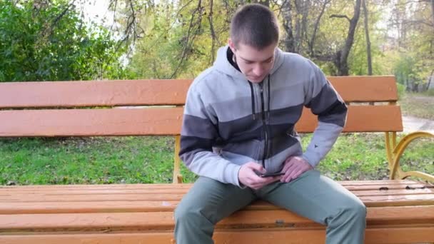 Молодой человек, сидящий на скамейке в парке с мобильным телефоном. Закрыть — стоковое видео