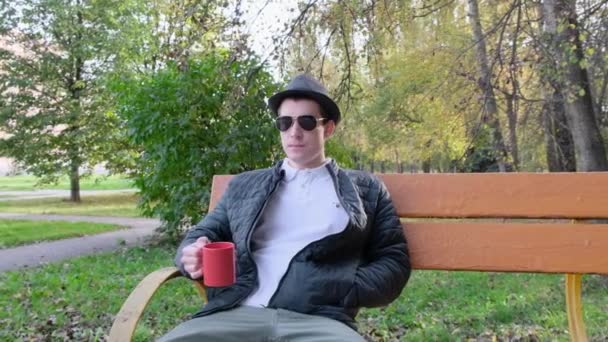 선글라스를 끼고 있는 젊은 남자가 벤치에서 유리 안에서 차를 마시고 있습니다. 선선 한 가을 날입니다. 닫아 — 비디오