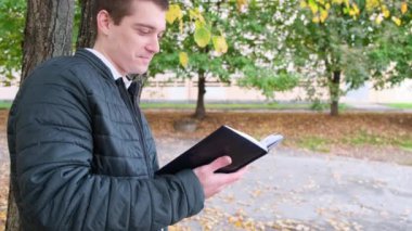 Geniş açılı genç adam parkta bir bankta kitap okuyor, boş zamanlarında parkta. Kapat.