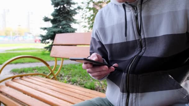 Un jeune homme assis sur un banc Park utilise l'écran tactile d'un smartphone. gros plan. — Video