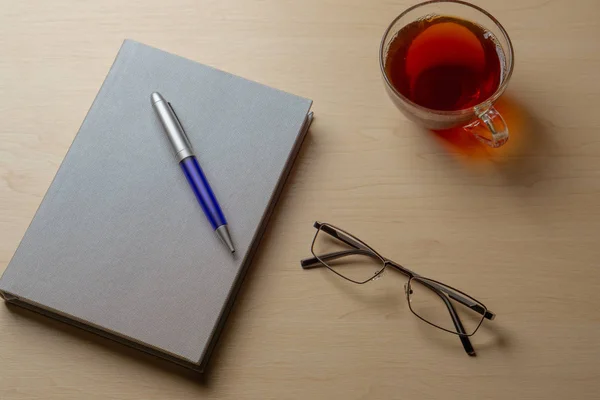 Gray diary, cup of tea, blue pen and glasses on a brown table, top view. Office desk table concept — Stock Photo, Image