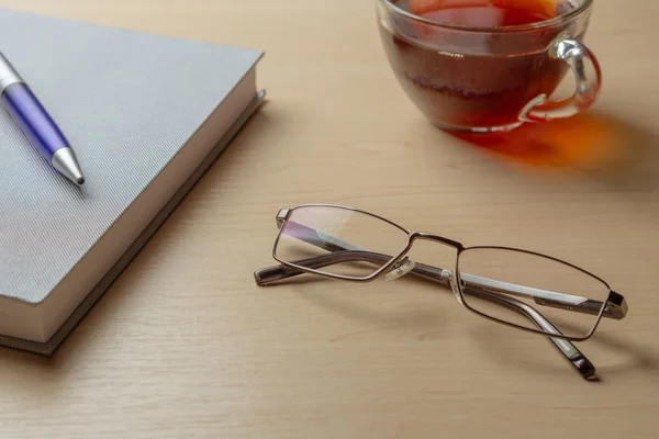 Eyeglasses, diary, blue pen and cup of tea on a brown table, selective focus — Stock Photo, Image