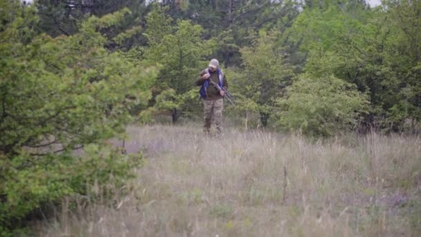 Um caçador com uma espingarda sai da floresta para uma clareira, um homem branco em camuflagem com uma espingarda — Vídeo de Stock
