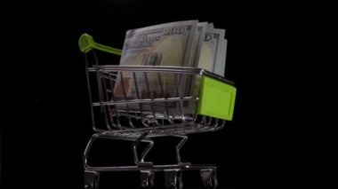 Bir alışveriş arabasında 100 dolarlık banknotlar, siyah bir arka planda dönüyor.
