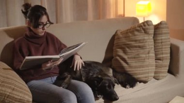 Bir kız büyük bir kitap okurken kanepede oturuyor. Bir kara köpeğin yanına kanepede da.