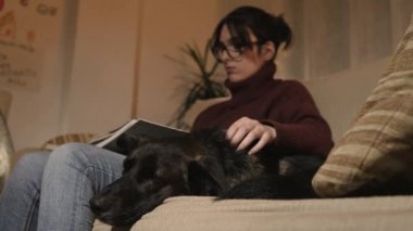 Bir kız büyük bir kitap okurken kanepede oturuyor. Bir kara köpeğin yanına kanepede da.