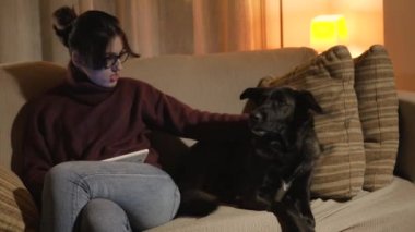 Bir kız kanepede oturan ve bir tablet tutan bir köpek ile oynuyor. Köpek de kanepede oturuyor.