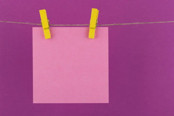 paper sheets of a notebook for notes and reminders of pink color fixed with decorative yellow clothespins hang on a rope on a lilac background