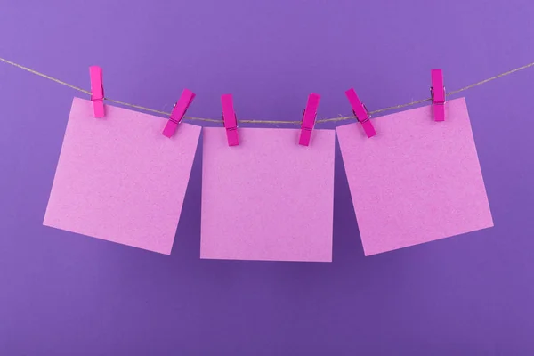 paper sheets of a notebook for notes and reminders of pink color fixed with decorative lilac clothespins hang on a rope on a lilac background