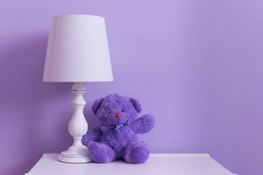 Beyaz bir masa lambası leylak duvara karşı bir oyuncak oyuncak ayı yanında beyaz bir komite üzerinde duruyor