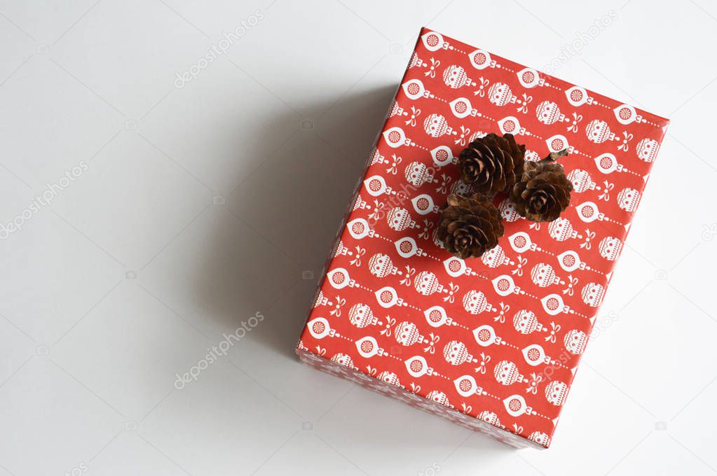 A holiday gift box. Holiday box for Christmas and New year. Red box with white pattern and fir cones on white background. Holiday gift. Preparation for the celebration.