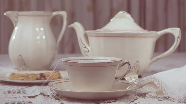 Table laid with delicate white thread tablecloth and elegant tea set. The dishes are porcelain and consist of a cup of tea, plates, teapot and milk. The cup contains a tea bag and water. One of the dishes has sweet cookies.