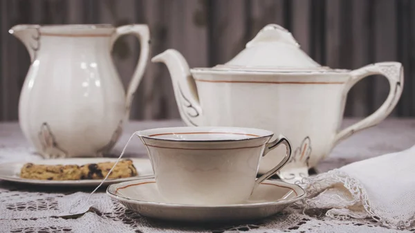 Table laid with delicate white thread tablecloth and elegant tea set. The dishes are porcelain and consist of a cup of tea, plates, teapot and milk. The cup contains a tea bag and water. One of the dishes has sweet cookies.