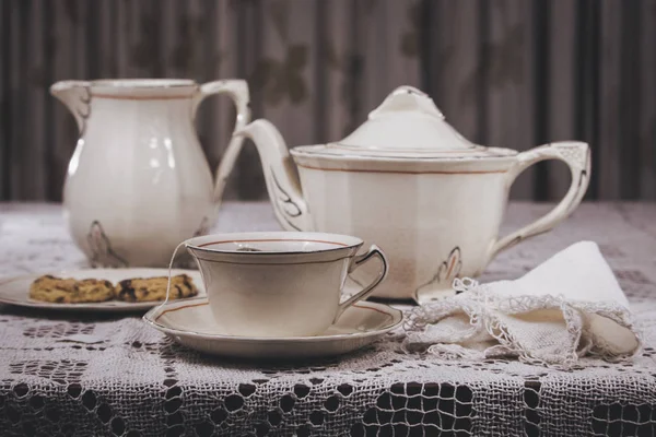 Table laid with delicate white thread tablecloth and elegant tea set. The dishes are porcelain and consist of a cup of tea, plates, teapot and milk. The cup contains a tea bag and water. One of the dishes has sweet cookies.