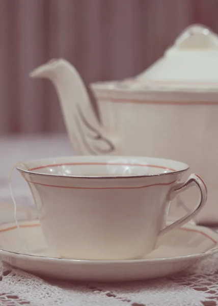 Table laid with delicate white thread tablecloth and elegant tea set. The dishes are porcelain and consist of a cup of tea, plates, teapot and milk. The cup contains a tea bag and water. One of the dishes has sweet cookies.