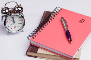 2020 yılından kalma, kırmızı, yeni ciltli gündem, üzerinde bir kalem olan. Aşağıda eski bir kitap var. Yan tarafında çanları olan antika gümüş bir saat var. Beyaz arkaplanda