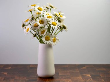 Beyaz vazo taze kesilmiş papatya beyaz ve sarı kır çiçekleriyle dolu kesme tahtası yüzeyinde beyaz bir duvarla oturuyor. Doğa, natürmort fotoğraf için içeri getirildi..