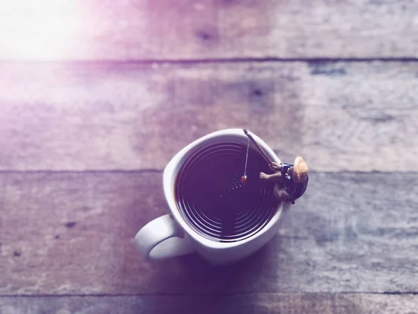 Relaxation in coffee time.Black coffee in white mug,Miniature of fisherman sitting and fishing, relaxed on the cup edge, in Americano coffee,free space for your text, with lens flare, selective focus.