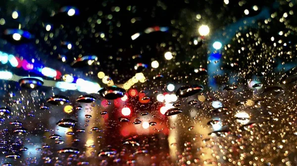 Defocused görüntü, gece sokak ışığı ile renkli bokeh, araba ön cam üzerinde yağmur damlası. Şiddetli yağmur fırtınasında araba sürmek. Yağmurlu bir günde şehirde trafik.