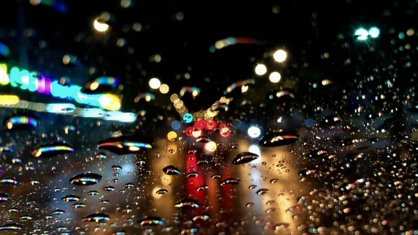 Defocused görüntü, gece sokak ışığı ile renkli bokeh, araba ön cam üzerinde yağmur damlası. Şiddetli yağmur fırtınasında araba sürmek. Yağmurlu bir günde şehirde trafik.