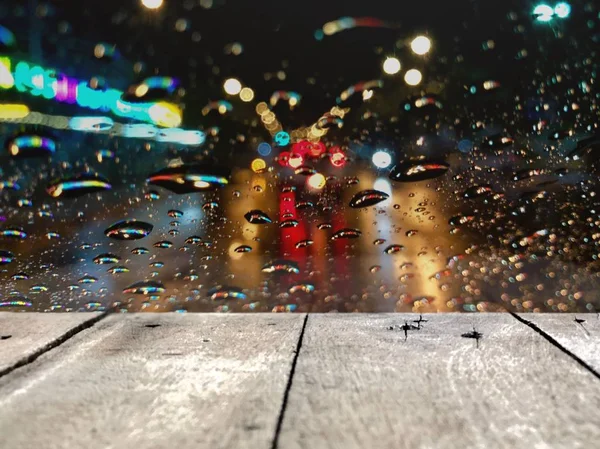 Perspective wood table top and blurred colorful bokeh with street light at night,raindrop on car windshield.Driving car in heavy rain storm.Traffic in the city on a rainy day,free space for your text