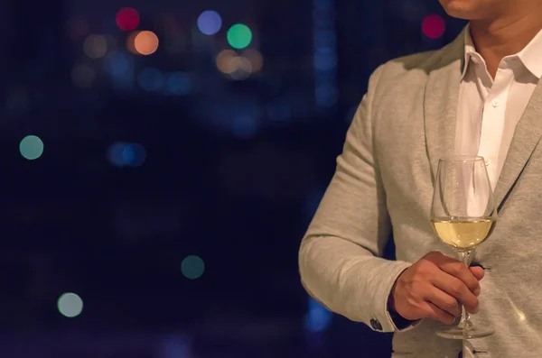 Businessman wearing grey color suit stands at the rooftop bar holding a glass of white wine with dark background of city bokeh lights.