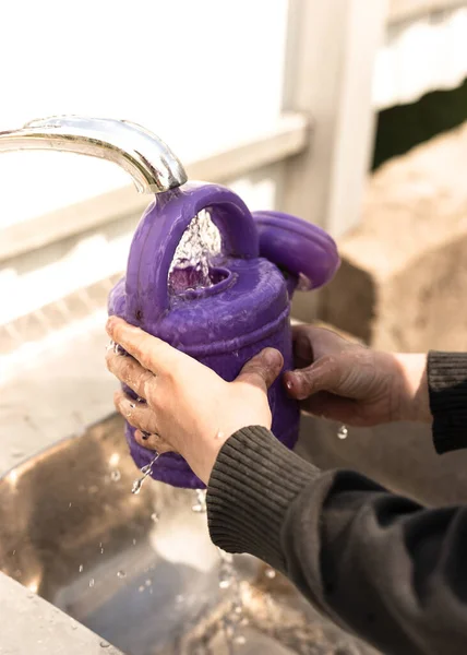 The boy pours water from a tap into a purple watering can