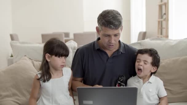Веселый папа и двое милых детей, сидящих на диване за ноутбуком — стоковое видео