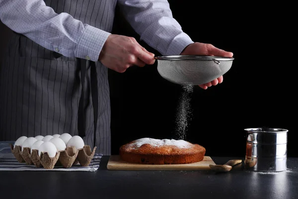 man powder of sugar to pie on dark background. decorating dessert concept with ingredients
